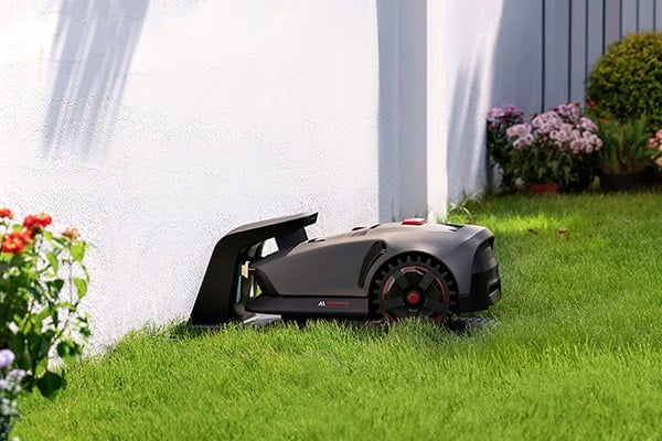 Na obrázku je sekací robot umístěný v nabíjecí stanici. Vlevo je vidět bílý dům a květiny.